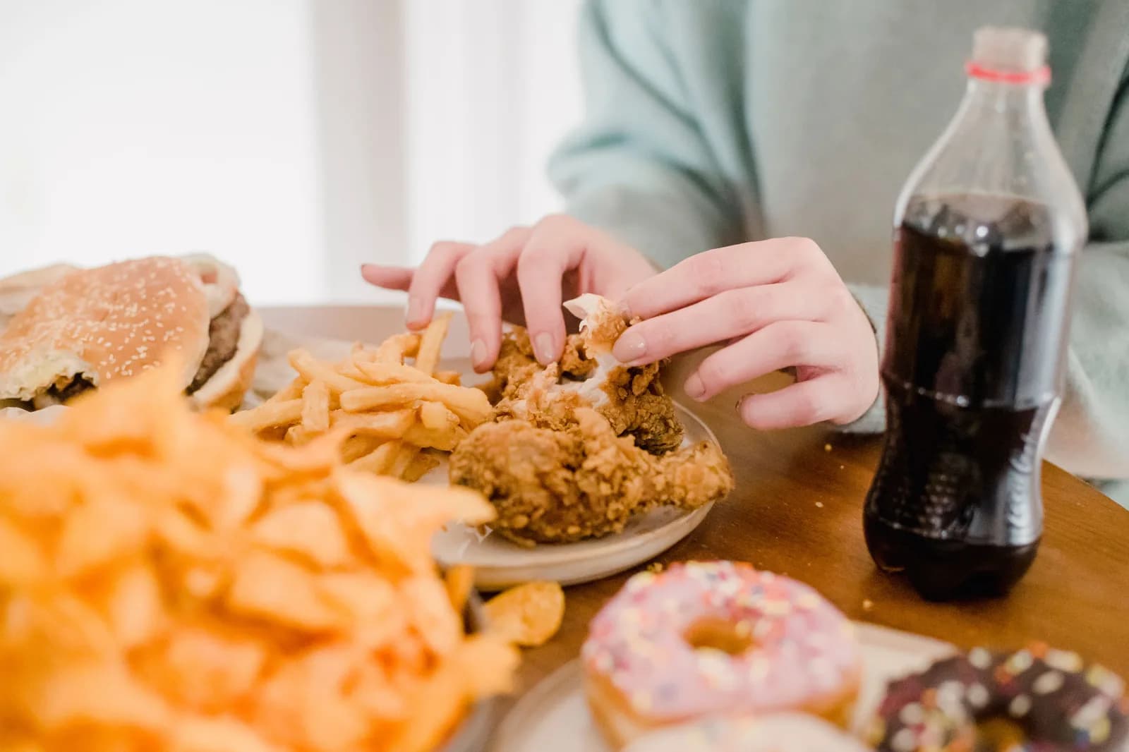 Person eating fried chicken at a table with fries, doughnuts and a soft drink