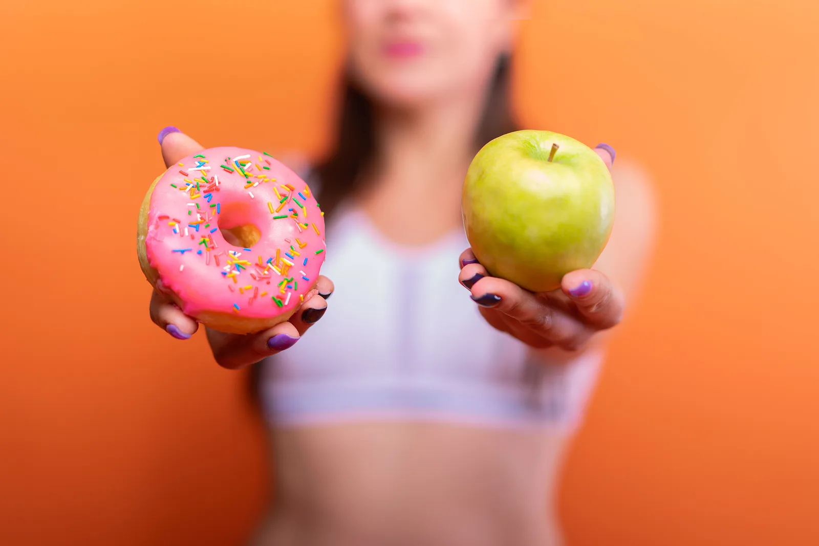 Woman holding a pink frosted doughnut in one hand and a green apple in the other